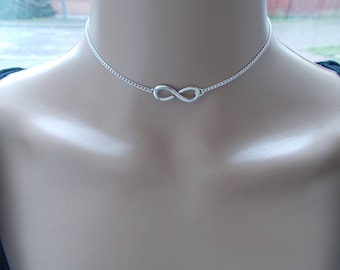 May include: A silver-colored choker necklace featuring an infinity symbol pendant. The necklace is made of a delicate chain and is worn around the neck. The infinity symbol is centered on the chest.