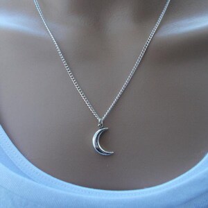 Pu&ograve; includere: Collana con pendente a forma di mezzaluna argentata. Il pendente ha una forma piccola e curva, sospesa a una delicata catena. La collana &egrave; indossata su un top bianco.