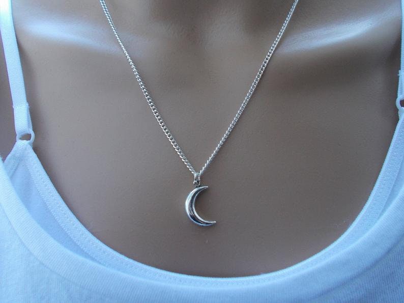 Pu&ograve; includere: Collana con pendente a forma di mezzaluna argentata. Il pendente ha una forma piccola e curva, sospesa a una delicata catena. La collana &egrave; indossata su un top bianco.