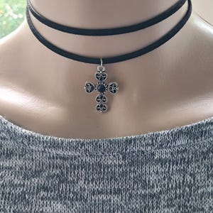 May include: A black choker necklace with a silver cross pendant. The cross is decorated with black gemstones and detailed patterns. The necklace has two black cords and is worn around the neck.