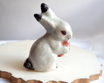 May include: A small ceramic rabbit figurine with gray fur, black ears, and a pink nose. The rabbit is sitting on a white surface with a gold rim.