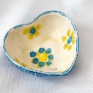 May include: A small heart-shaped ceramic dish with a blue rim and white interior. The dish is decorated with yellow and blue flowers.