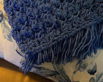 Peut inclure: Une couverture en crochet bleu avec des franges. La couverture est posée sur un oreiller blanc à motif floral.