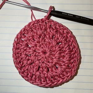 May include: A close-up of a pink crocheted circle with a textured pattern. The circle is being worked on with a crochet hook and yarn.