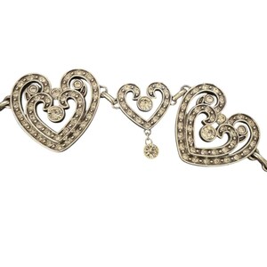 May include: A silver-toned metal necklace featuring three heart-shaped links. The two larger outer hearts have intricate scrollwork and are encrusted with small clear rhinestones. A smaller central heart features a dangling rhinestone charm.