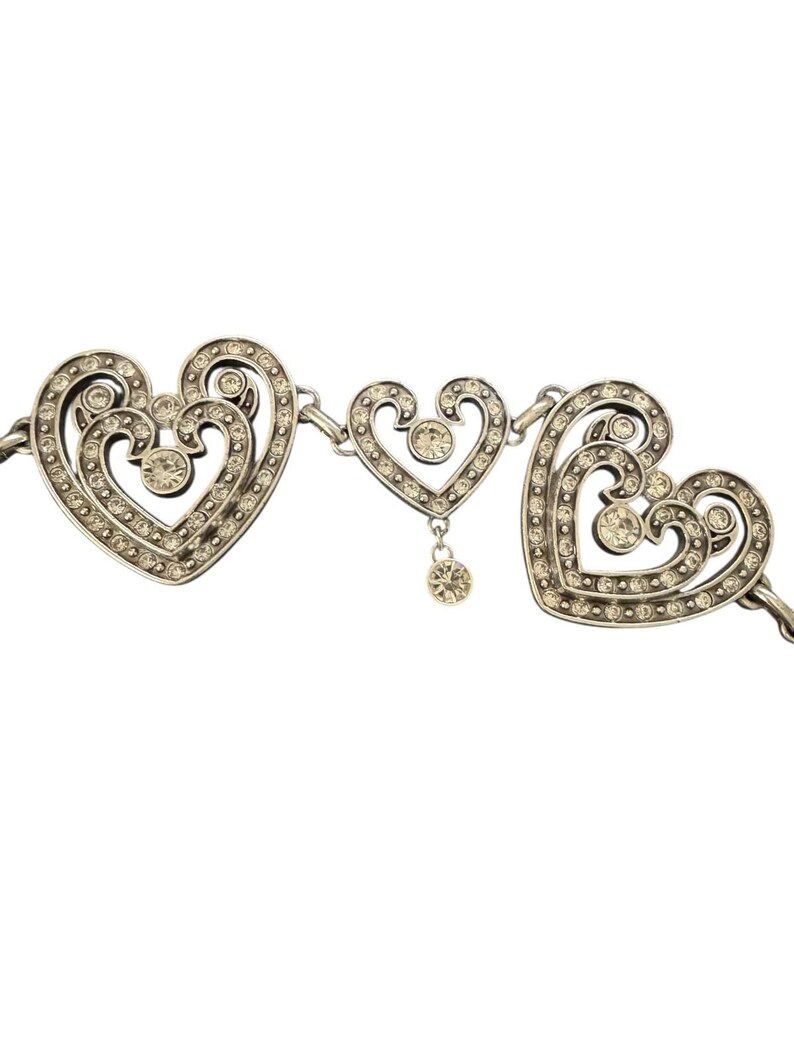 May include: A silver-toned metal necklace featuring three heart-shaped links. The two larger outer hearts have intricate scrollwork and are encrusted with small clear rhinestones. A smaller central heart features a dangling rhinestone charm.