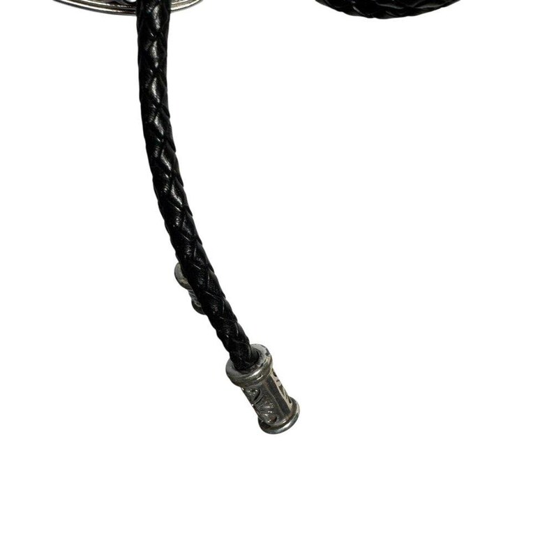 K&ouml;nnte beinhalten: Nahaufnahme einer schwarzen geflochtenen Leder-Bolo-Krawatte. Die Krawatte hat einen silberfarbenen dekorativen Verschluss am Ende mit eingravierten Details. Die Bolo-Krawatte ist ein klassisches Accessoire.