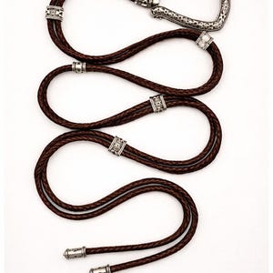 Puede incluir: Una corbata bolo de cuero marr&oacute;n con una hebilla y detalles decorativos plateados. El cord&oacute;n de cuero trenzado est&aacute; en bucle y presenta detalles met&aacute;licos ornamentados. Los extremos de la corbata est&aacute;n acabados con puntas plateadas a juego.