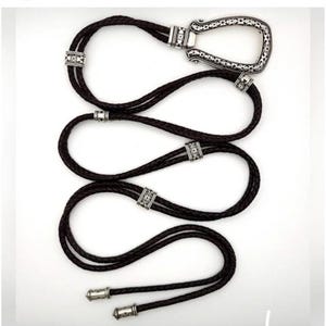 K&ouml;nnte beinhalten: Dunkelbraune geflochtene Leder-Bolo-Krawatte mit dekorativem silberfarbenem Verschluss und Akzenten. Der Verschluss ist ein gro&szlig;es, verziertes Rechteck. Die Krawatte hat drei Str&auml;nge aus geflochtenem Leder und silberfarbene Metalldetails.