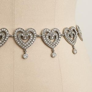 May include: A silver-toned metal chain belt featuring a series of repeating heart-shaped links. Each heart is encrusted with small clear rhinestones and includes a single dangling circular rhinestone charm suspended from the bottom center point.