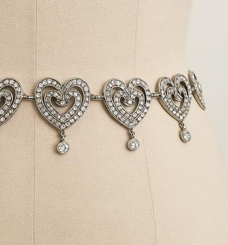 May include: A silver-toned metal chain belt featuring a series of repeating heart-shaped links. Each heart is encrusted with small clear rhinestones and includes a single dangling circular rhinestone charm suspended from the bottom center point.