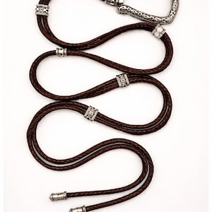 Puede incluir: Una corbata bolo de cuero marr&oacute;n con una hebilla y detalles decorativos plateados. El cord&oacute;n de cuero trenzado est&aacute; en bucle y presenta detalles met&aacute;licos ornamentados. Los extremos de la corbata est&aacute;n acabados con puntas plateadas a juego.