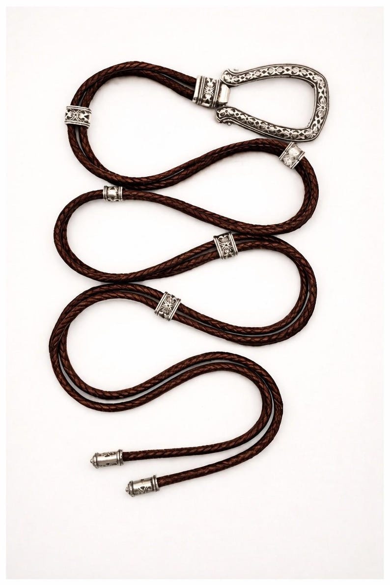 Puede incluir: Una corbata bolo de cuero marr&oacute;n con una hebilla y detalles decorativos plateados. El cord&oacute;n de cuero trenzado est&aacute; en bucle y presenta detalles met&aacute;licos ornamentados. Los extremos de la corbata est&aacute;n acabados con puntas plateadas a juego.