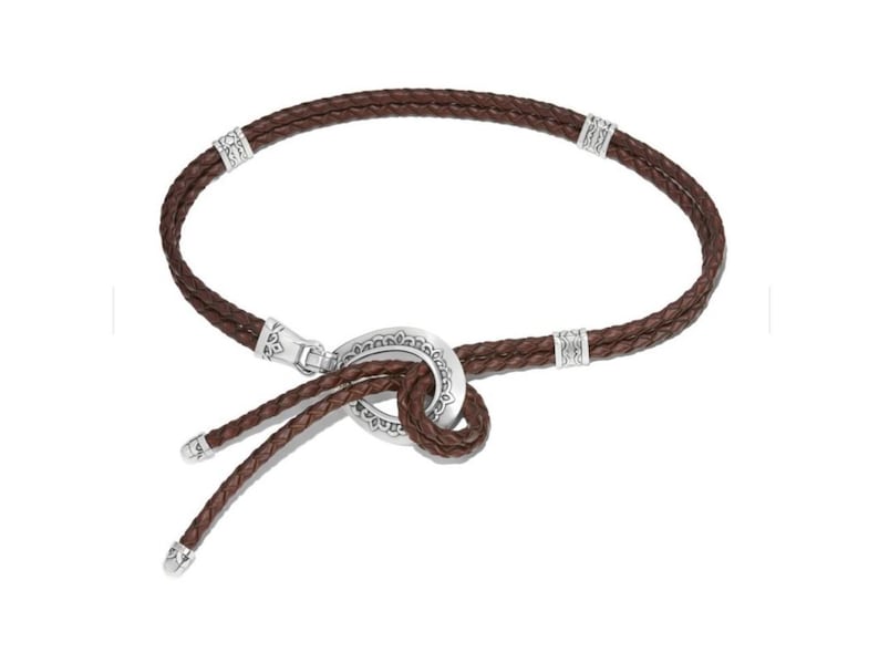 May include: A brown leather necklace with a braided design. The necklace has silver-coloured metal accents and a decorative clasp. The design includes a knot-like feature in the centre, with two braided leather strands hanging down.