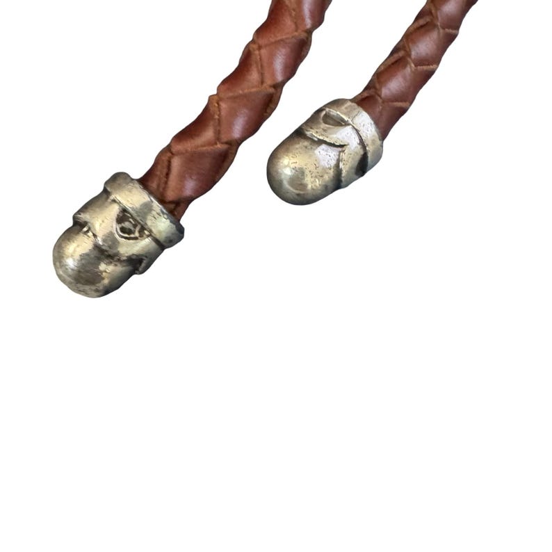 May include: Close-up of a brown leather braided cord with silver-coloured skull-shaped end caps. The cord is a rich, warm brown colour, and the metal caps have a detailed skull design. The background is plain white.