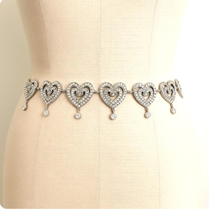 May include: A silver-toned metal waist chain featuring a series of repeating heart-shaped links. Each heart is embellished with small clear stones and includes a single dangling circular stone charm suspended from the bottom center of each link.