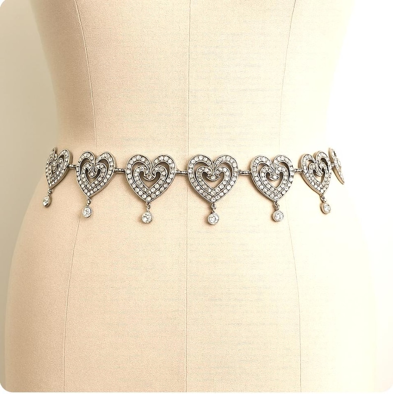 May include: A silver-toned metal waist chain featuring a series of repeating heart-shaped links. Each heart is embellished with small clear stones and includes a single dangling circular stone charm suspended from the bottom center of each link.