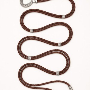 May include: A brown leather belt with a silver buckle and decorative accents. The braided belt has multiple silver embellishments. The buckle is a unique teardrop shape. The belt's length is approximately 100 cm.