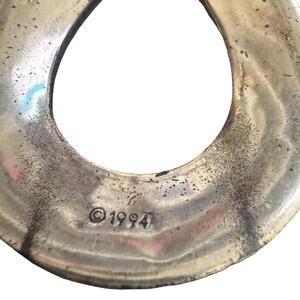 May include: Close-up of a silver-toned, abstract metal object with a loop shape. The surface has a textured, aged appearance. The year "&copy;1994" is stamped on the metal. The object is set against a white background.
