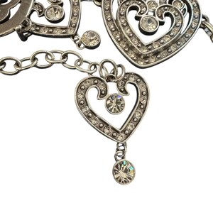 May include: A silver-toned metal chain belt featuring multiple heart-shaped links. Each heart is embellished with small, clear, round rhinestones set along the edges and a single larger rhinestone at the center. A dangling heart charm hangs from the chain.