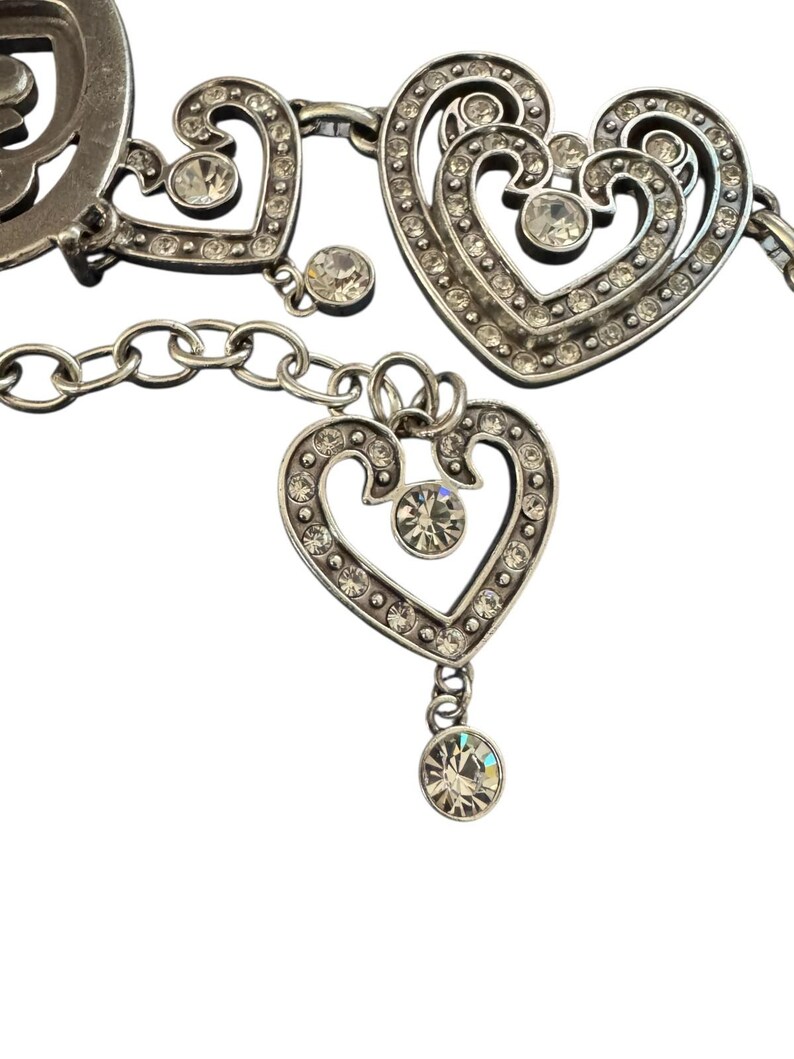 May include: A silver-toned metal chain belt featuring multiple heart-shaped links. Each heart is embellished with small, clear, round rhinestones set along the edges and a single larger rhinestone at the center. A dangling heart charm hangs from the chain.