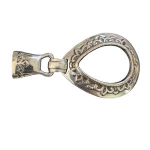 May include: A silver-coloured metal belt buckle with an ornate teardrop-shaped design. The buckle features a decorative floral pattern around the teardrop's edge and a clasp mechanism on one end. The buckle is designed to be used with a belt.