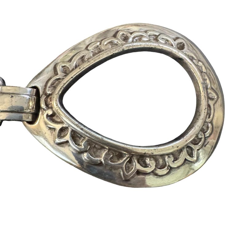 May include: A silver-coloured metal belt buckle with an oval shape and decorative floral detailing around the edges. The buckle has a hinged mechanism on one side for attachment to a belt.