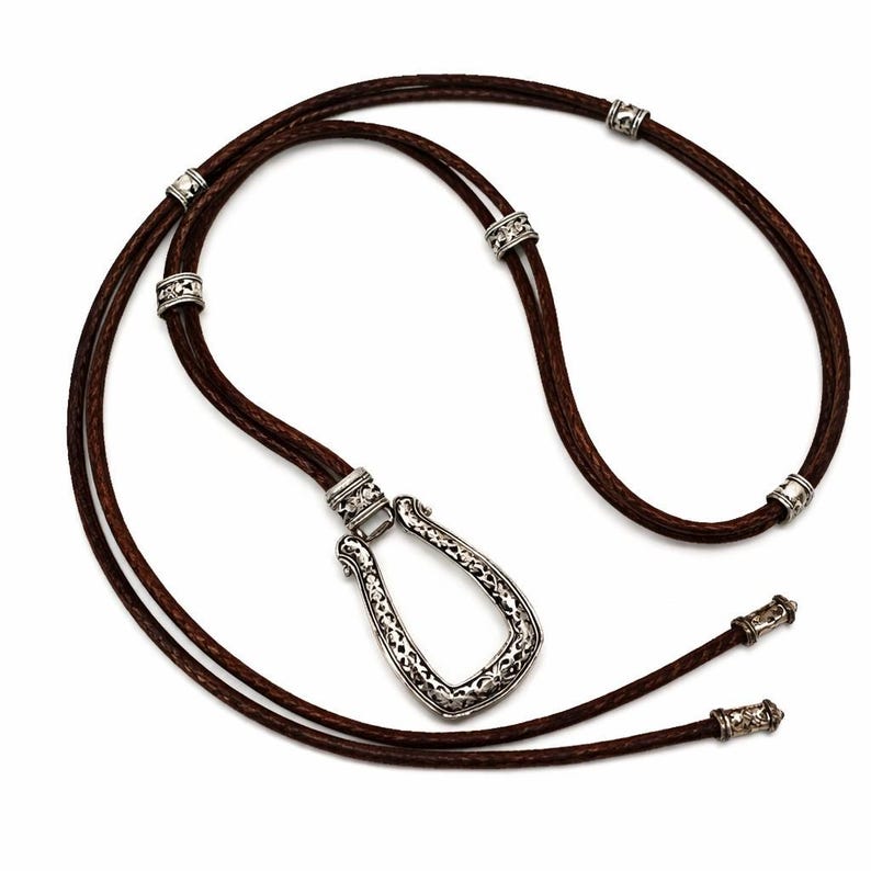 Puede incluir: Un collar de cuero marr&oacute;n con un colgante plateado en forma de herradura. El colgante tiene un dise&ntilde;o calado y ornamentado. El collar tambi&eacute;n presenta cuentas y extremos decorativos plateados.