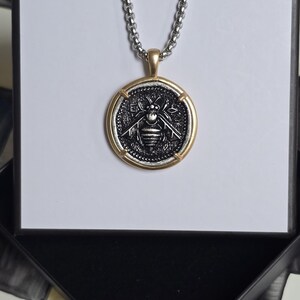 Peut inclure: Un collier &agrave; cha&icirc;ne argent&eacute;e avec un pendentif rond orn&eacute; d'un motif d'abeille en relief. Le pendentif pr&eacute;sente un centre sombre et textur&eacute; avec un dessin d'abeille, encadr&eacute; par une bordure dor&eacute;e lisse. Le collier est pr&eacute;sent&eacute; sur un support blanc.