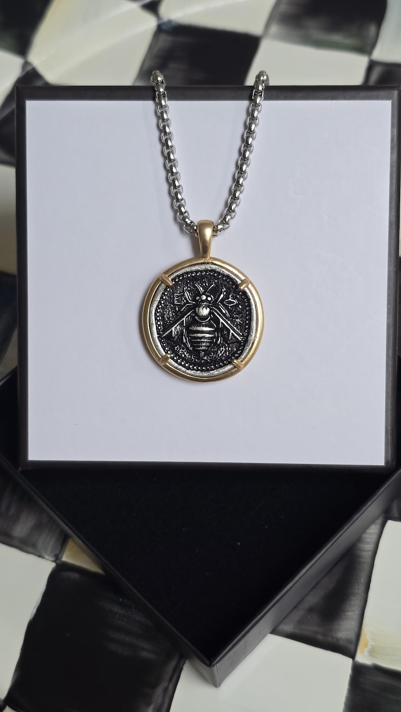 Peut inclure: Un collier &agrave; cha&icirc;ne argent&eacute;e avec un pendentif rond orn&eacute; d'un motif d'abeille en relief. Le pendentif pr&eacute;sente un centre sombre et textur&eacute; avec un dessin d'abeille, encadr&eacute; par une bordure dor&eacute;e lisse. Le collier est pr&eacute;sent&eacute; sur un support blanc.