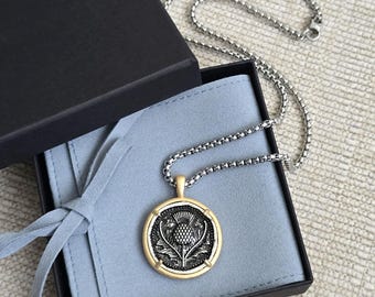 Könnte beinhalten: Halskette mit silberner Kette und rundem Anhänger mit Distel-Design. Der Anhänger hat einen goldfarbenen Rand und ein detailliertes silbernes Distel-Motiv. Die Halskette wird in einer schwarzen Geschenkbox mit hellblauem Innenraum präsentiert.