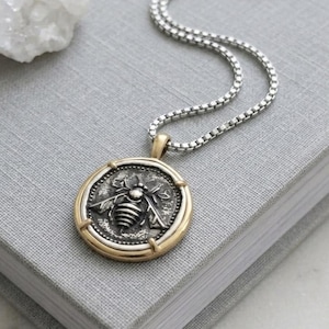 Peut inclure: Un collier &agrave; cha&icirc;ne carr&eacute;e en argent avec un pendentif rond orn&eacute; d'un motif d'abeille en relief. Le pendentif pr&eacute;sente un centre en argent fonc&eacute; avec une abeille, entour&eacute; d'un bord en m&eacute;tal dor&eacute; lisse. Le collier est pos&eacute; sur une surface grise.