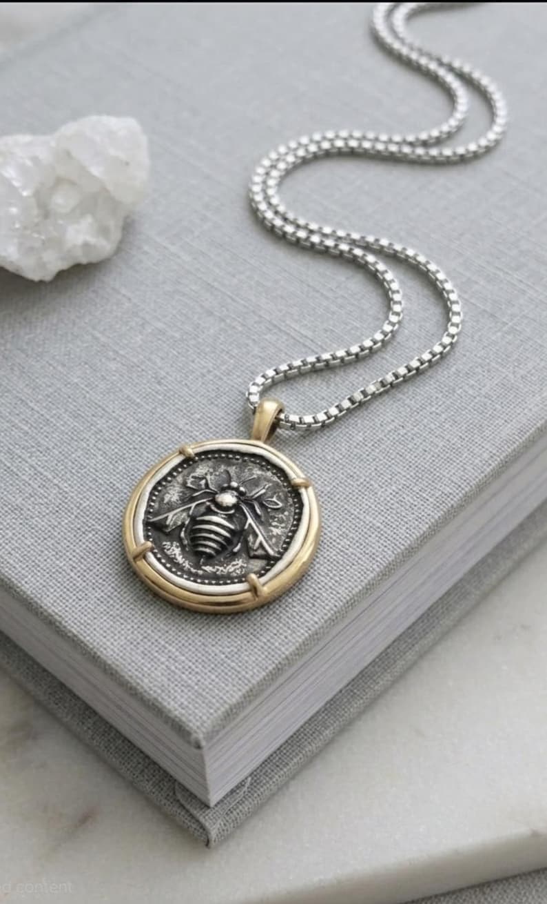 Peut inclure: Un collier &agrave; cha&icirc;ne carr&eacute;e en argent avec un pendentif rond orn&eacute; d'un motif d'abeille en relief. Le pendentif pr&eacute;sente un centre en argent fonc&eacute; avec une abeille, entour&eacute; d'un bord en m&eacute;tal dor&eacute; lisse. Le collier est pos&eacute; sur une surface grise.