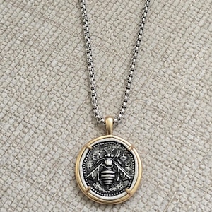 Peut inclure: Un collier &agrave; cha&icirc;ne maille carr&eacute;e argent&eacute;e avec un pendentif rond. Le pendentif pr&eacute;sente un relief d'abeille argent&eacute; au centre sur fond sombre, encadr&eacute; par un bord circulaire dor&eacute; lisse avec quatre petits accents dor&eacute;s sur les bords.
