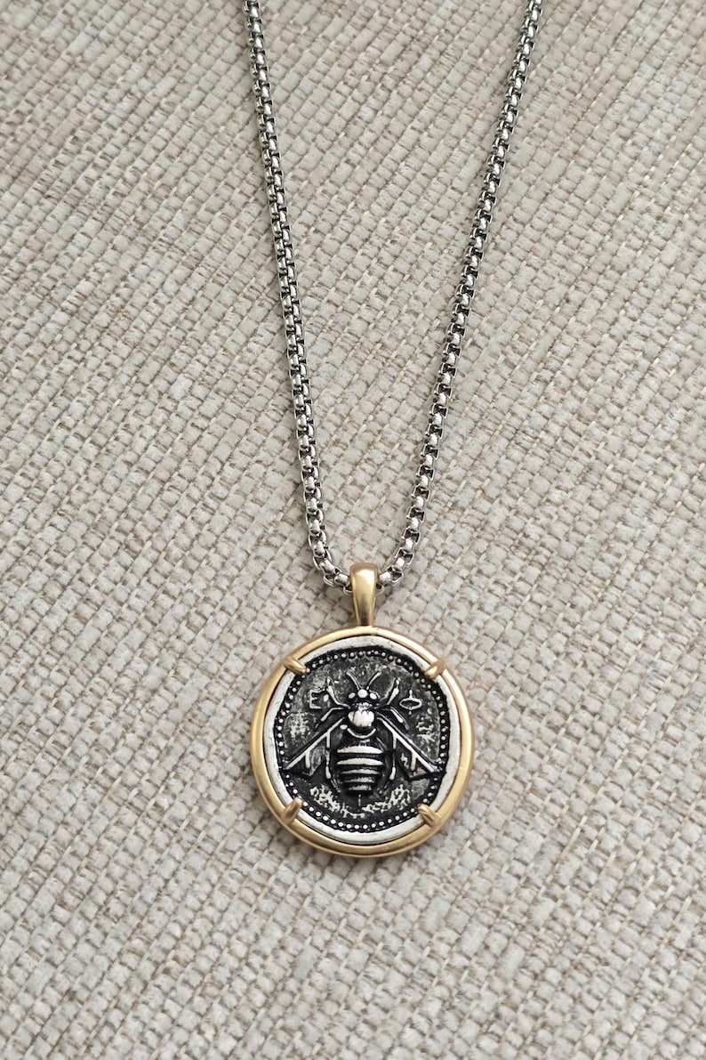 Peut inclure: Un collier &agrave; cha&icirc;ne maille carr&eacute;e argent&eacute;e avec un pendentif rond. Le pendentif pr&eacute;sente un relief d'abeille argent&eacute; au centre sur fond sombre, encadr&eacute; par un bord circulaire dor&eacute; lisse avec quatre petits accents dor&eacute;s sur les bords.
