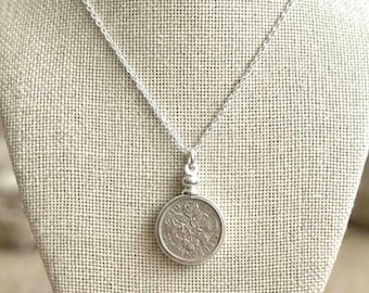 Peut inclure: Un collier pendentif argenté avec un pendentif pièce de monnaie. La pièce, ornée de détails complexes, est suspendue à une fine chaîne. Le collier est présenté sur un présentoir de couleur neutre.