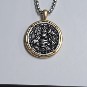 Peut inclure: Un collier pendentif rond compos&eacute; d'une pi&egrave;ce argent&eacute;e avec un relief d'abeille d&eacute;taill&eacute; au centre, encadr&eacute;e par un contour en m&eacute;tal dor&eacute; lisse. Le pendentif est suspendu &agrave; une cha&icirc;ne &agrave; billes argent&eacute;e, cr&eacute;ant un contraste entre les finitions.