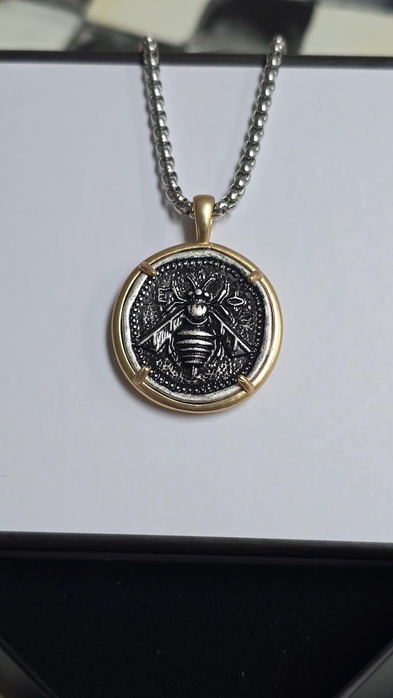 Peut inclure: Un collier pendentif rond compos&eacute; d'une pi&egrave;ce argent&eacute;e avec un relief d'abeille d&eacute;taill&eacute; au centre, encadr&eacute;e par un contour en m&eacute;tal dor&eacute; lisse. Le pendentif est suspendu &agrave; une cha&icirc;ne &agrave; billes argent&eacute;e, cr&eacute;ant un contraste entre les finitions.