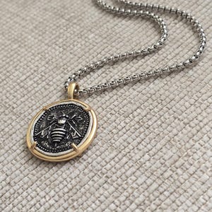 Peut inclure: Un collier &agrave; cha&icirc;ne carr&eacute;e argent&eacute;e avec un pendentif rond dot&eacute; d'un bord dor&eacute;. Le pendentif pr&eacute;sente un centre sombre et textur&eacute; avec un motif d'abeille argent&eacute; en relief et les lettres E et A de chaque c&ocirc;t&eacute; de la t&ecirc;te de l'insecte.