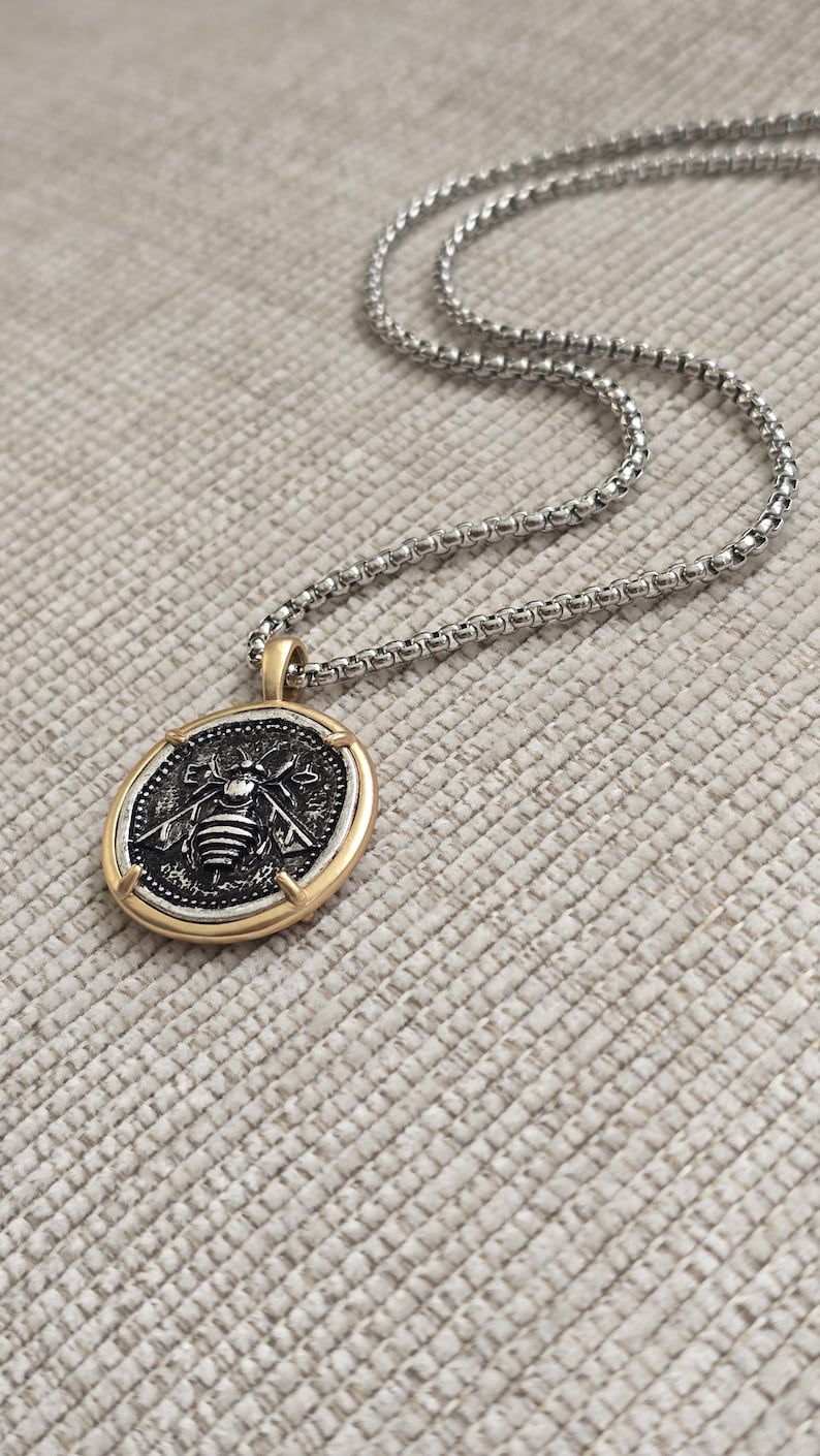 Peut inclure: Un collier &agrave; cha&icirc;ne carr&eacute;e argent&eacute;e avec un pendentif rond dot&eacute; d'un bord dor&eacute;. Le pendentif pr&eacute;sente un centre sombre et textur&eacute; avec un motif d'abeille argent&eacute; en relief et les lettres E et A de chaque c&ocirc;t&eacute; de la t&ecirc;te de l'insecte.