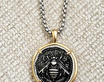 Peut inclure: Un collier argenté avec un pendentif doré. Le pendentif présente un motif d'abeille détaillé dans un cadre circulaire. La chaîne est de style maille carrée. Le fond est un tissu texturé de couleur claire.