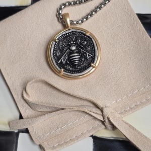 Peut inclure: Un collier pendentif rond compos&eacute; d'une pi&egrave;ce argent&eacute;e avec un relief d'abeille d&eacute;taill&eacute; au centre, encadr&eacute;e par un cercle en m&eacute;tal dor&eacute; lisse. Le pendentif est suspendu &agrave; une cha&icirc;ne &agrave; billes argent&eacute;e sur une pochette en tissu beige clair.