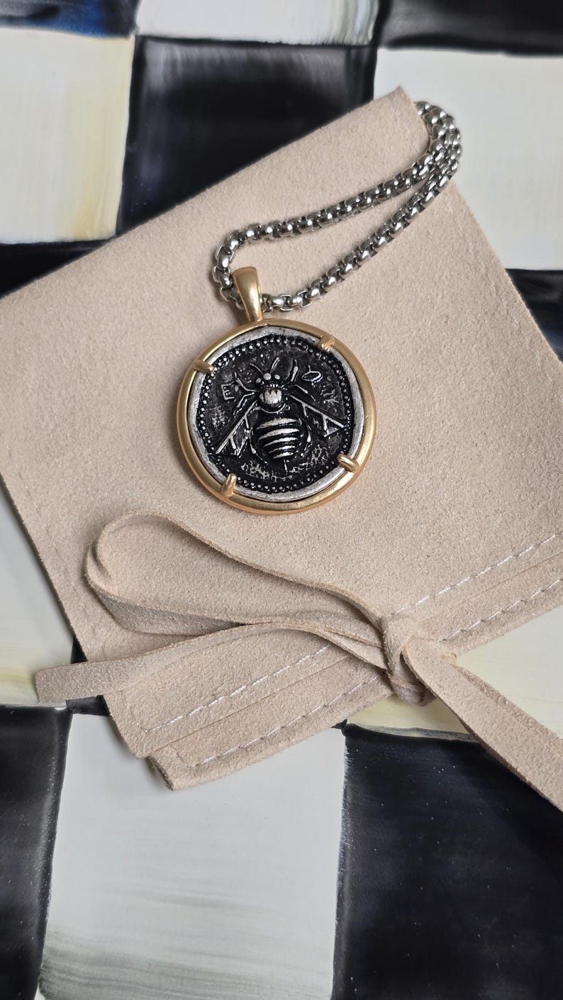 Peut inclure: Un collier pendentif rond compos&eacute; d'une pi&egrave;ce argent&eacute;e avec un relief d'abeille d&eacute;taill&eacute; au centre, encadr&eacute;e par un cercle en m&eacute;tal dor&eacute; lisse. Le pendentif est suspendu &agrave; une cha&icirc;ne &agrave; billes argent&eacute;e sur une pochette en tissu beige clair.