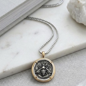 Peut inclure: Un collier &agrave; cha&icirc;ne bo&icirc;te argent&eacute;e avec un pendentif circulaire. Le pendentif pr&eacute;sente un centre en argent oxyd&eacute; fonc&eacute; grav&eacute; d'un motif d'abeille, encadr&eacute; par une bordure en m&eacute;tal dor&eacute; poli avec quatre petites griffes maintenant la pi&egrave;ce.
