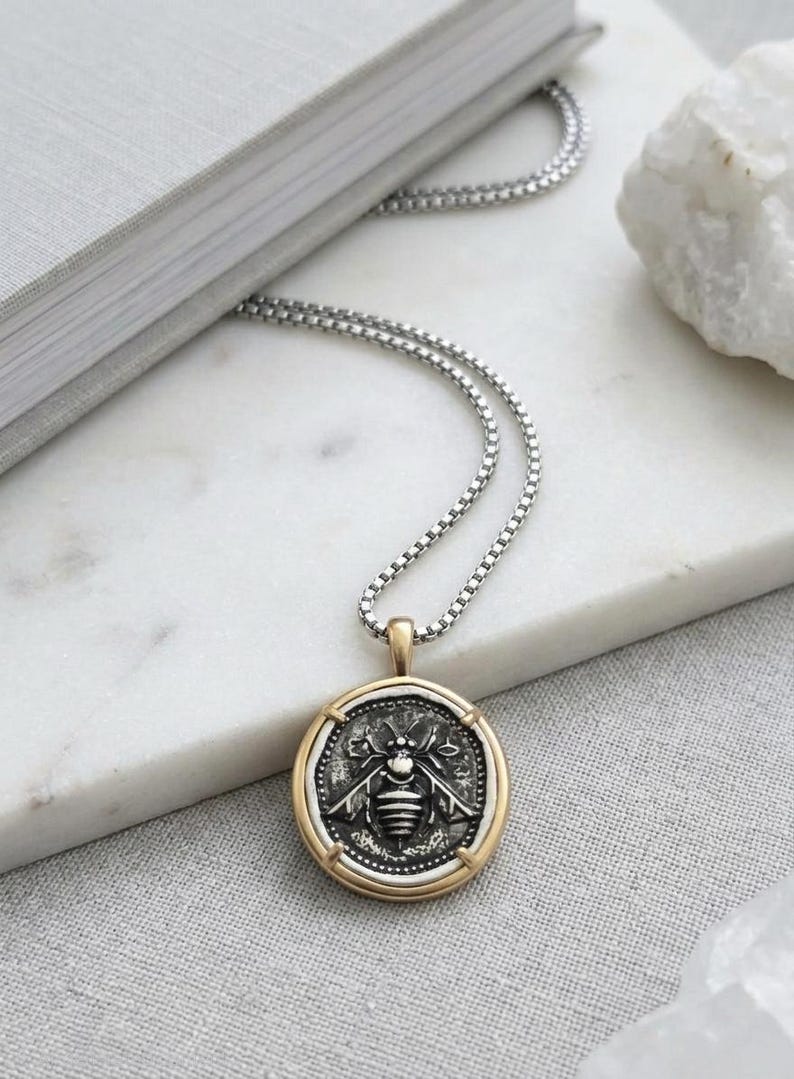 Peut inclure: Un collier &agrave; cha&icirc;ne bo&icirc;te argent&eacute;e avec un pendentif circulaire. Le pendentif pr&eacute;sente un centre en argent oxyd&eacute; fonc&eacute; grav&eacute; d'un motif d'abeille, encadr&eacute; par une bordure en m&eacute;tal dor&eacute; poli avec quatre petites griffes maintenant la pi&egrave;ce.
