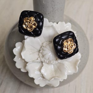 May include: A pair of black square earrings with a quilted design and a gold-colored flower in the center. The earrings are displayed on a white floral-shaped object, with a gray vase in the background.