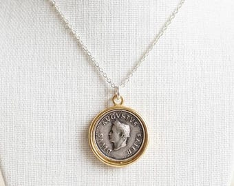 Peut inclure: Un collier en argent avec un pendentif pièce de monnaie dans une lunette dorée. La pièce de monnaie représente un profil et du texte. Le collier est présenté sur un buste blanc.