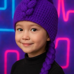 May include: A child wearing a vibrant purple knitted hat with a braided side. The hat is a deep violet color, and the braid hangs down the side. The child is wearing a black turtleneck. The background has neon lights.