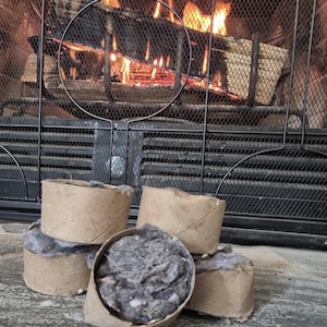May include: A collection of firelighters made from recycled materials. Each firelighter is a cardboard cylinder filled with a grey, fibrous substance. A fireplace with burning logs is visible in the background.