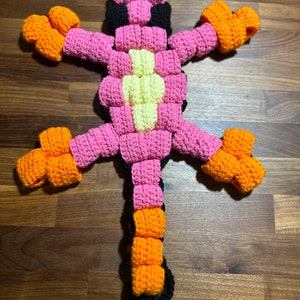 May include: A crocheted lizard toy with a pink, orange, yellow and black colour scheme. The lizard has a long tail and four legs.